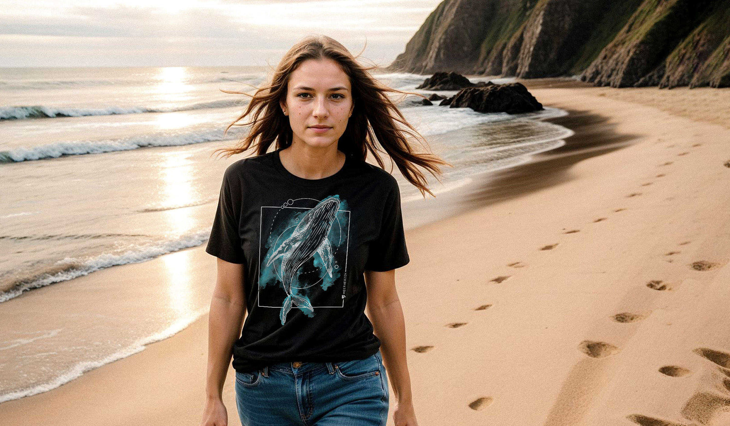 Eine Frau mit langen Haaren trägt ein schwarzes T-Shirt mit Wal-Motiv und spaziert am Strand entlang.