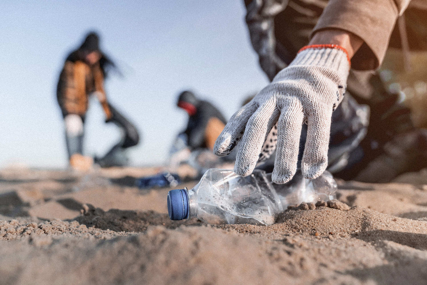 Hand greift am Strand nach einer Plastikflasche im Sand.